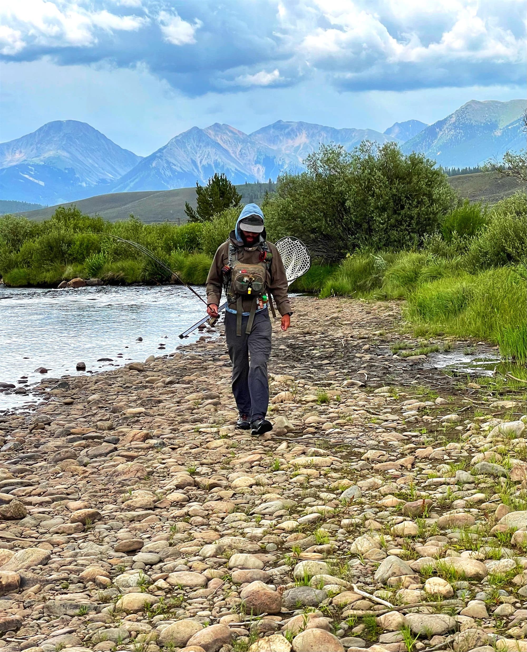 A fly fishing guide walking a river bank with a net, mountains behind him